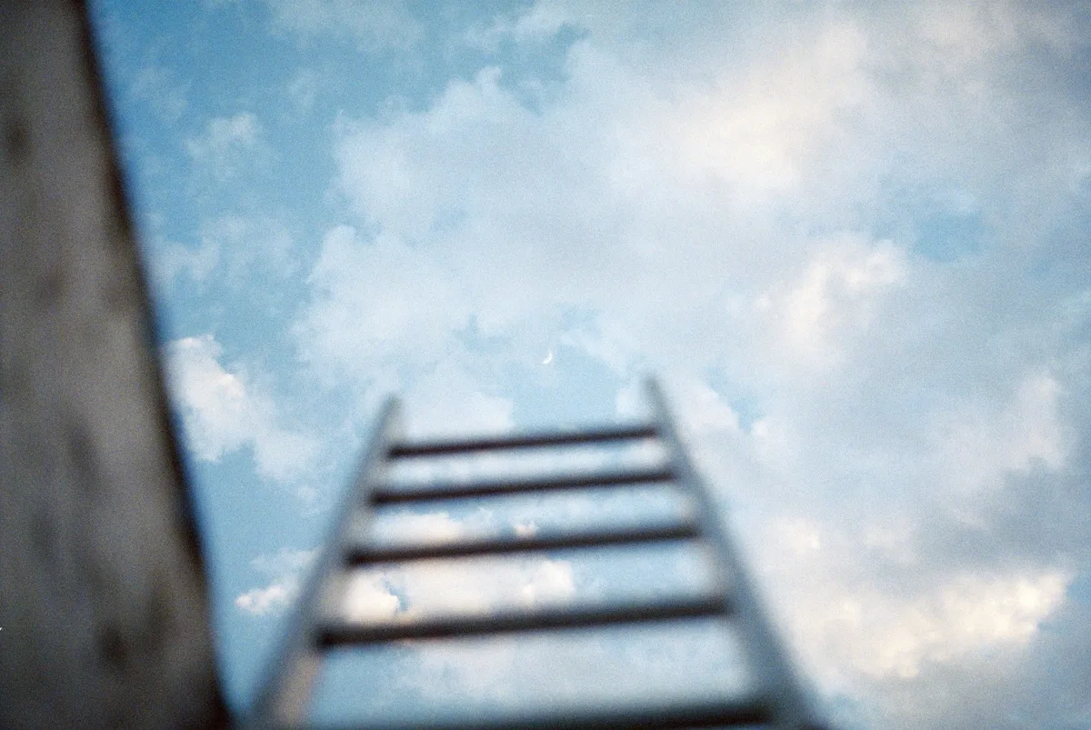 A ladder leaning against a wall, rising into a bright cloudy sky