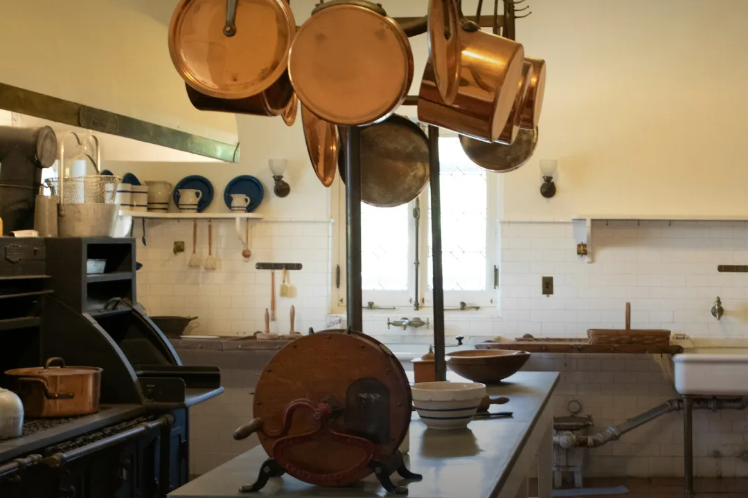 A grand old service kitchen with copper pans hanging above a steel-topped island and a wooden board standing on end